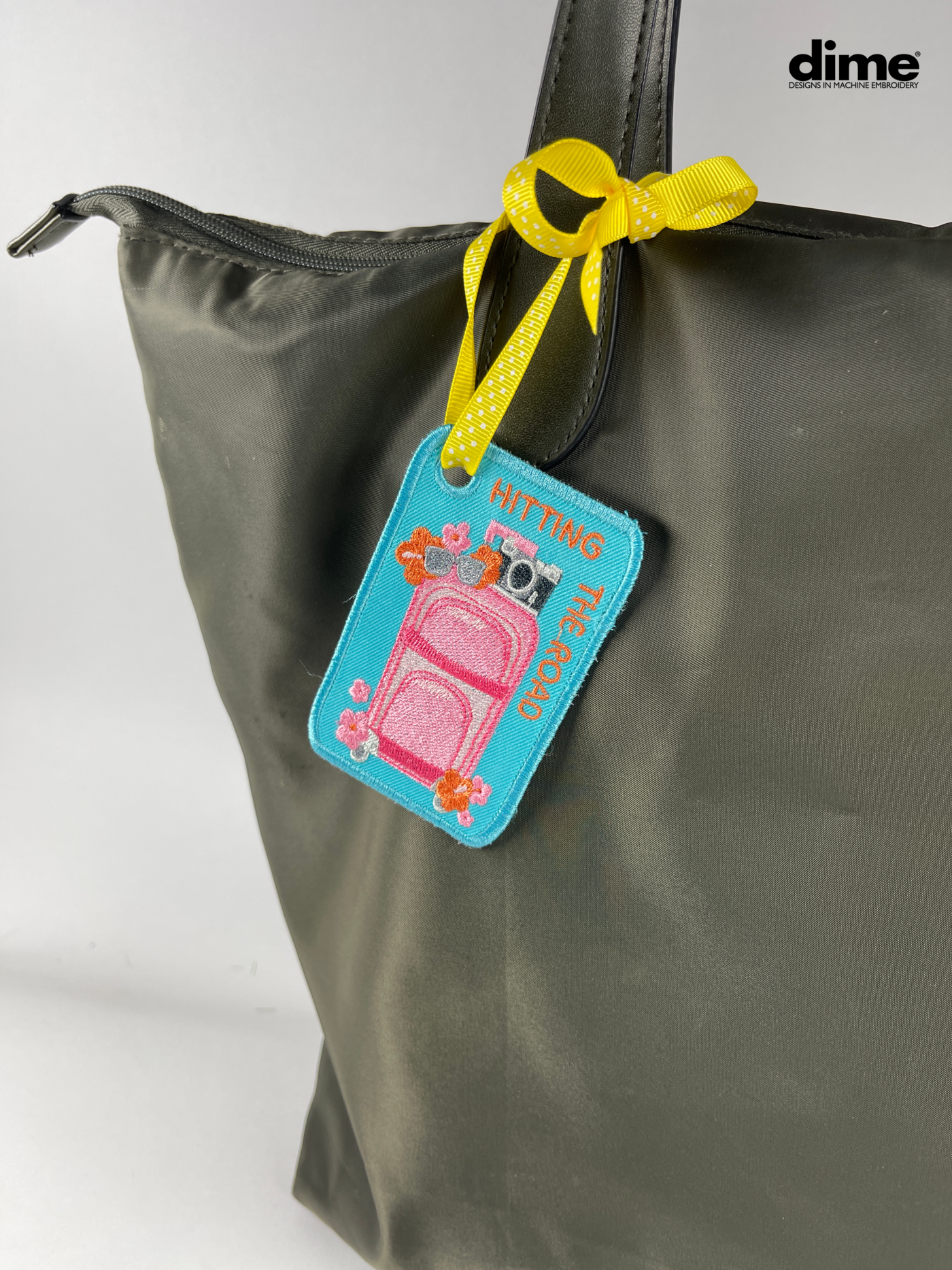 Tripping Luggage Tag n blue and pink attached to a grey bag with yellow ribbon